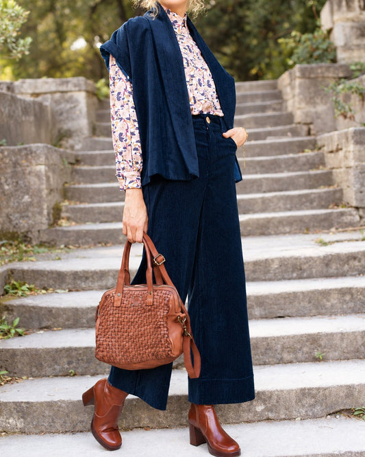 Pantalon Marinette en velours côtelé bio marine porté avec une chemise fleurie, une veste sans manches et un sac marron, en extérieur.