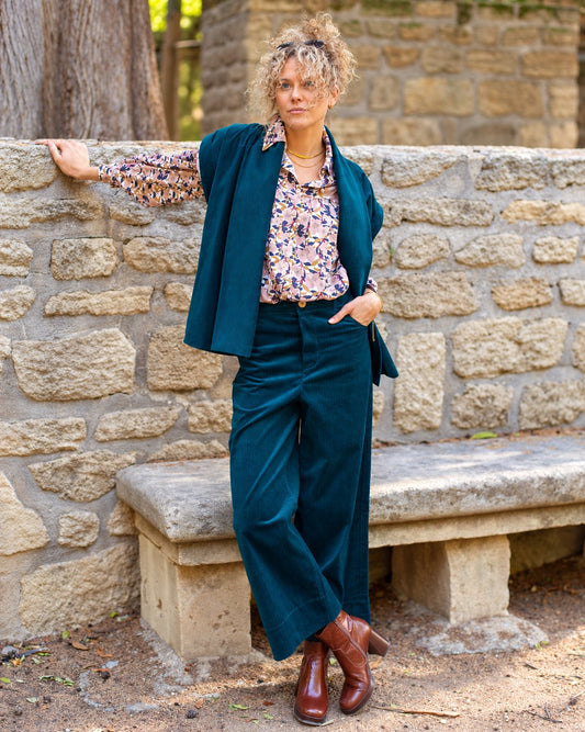 Pantalon Marinette en velours côtelé bio canard porté en pied avec une chemise fleurie et une veste assortie, devant un mur en pierre.
